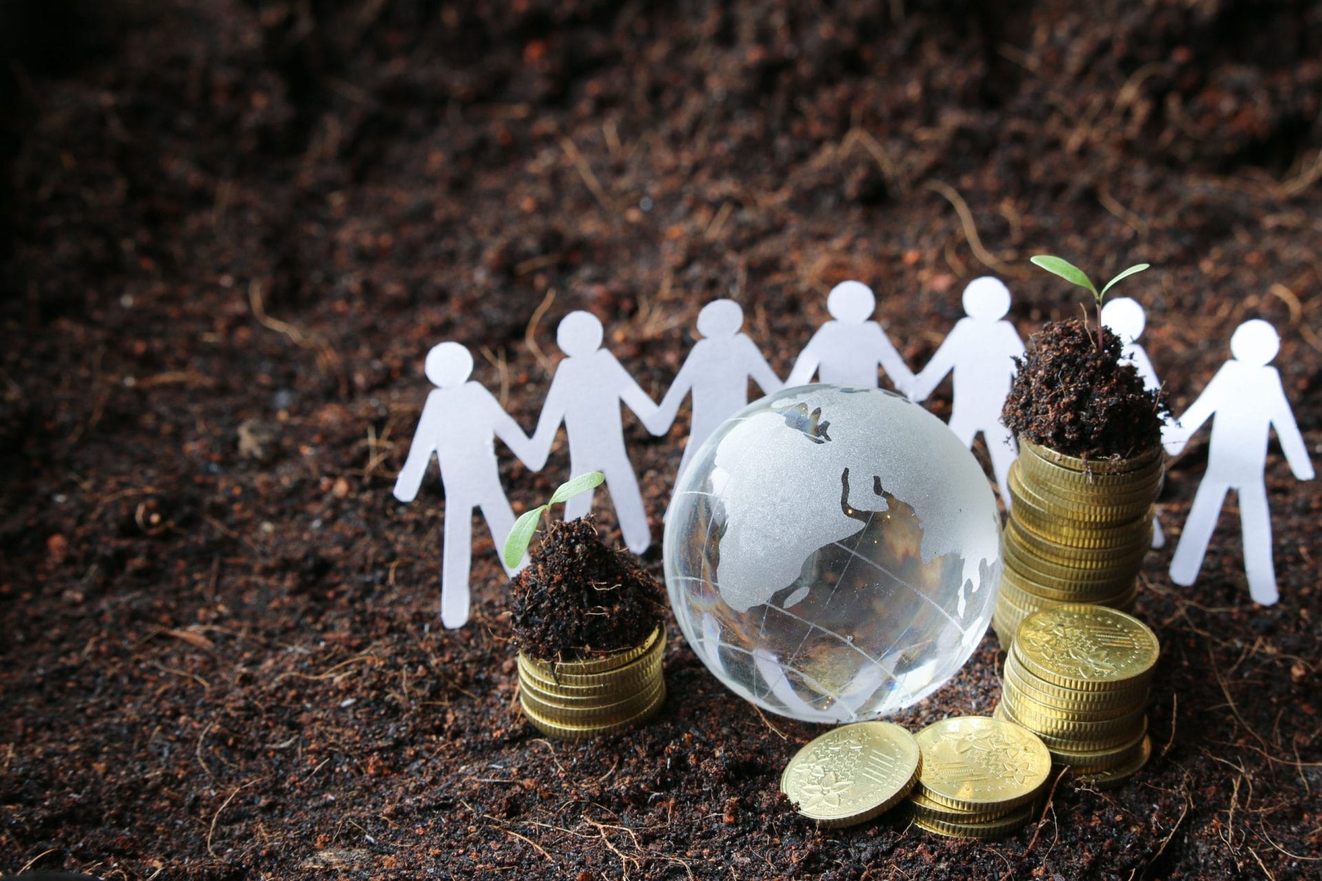 Aufnahme einer Menschenkette aus Papier, die vor einem kleinem Globus stehen und einem Münzhaufen